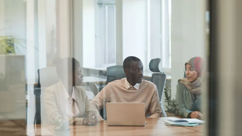 Three colleagues in discussion around a laptop in a glass-walled meeting room