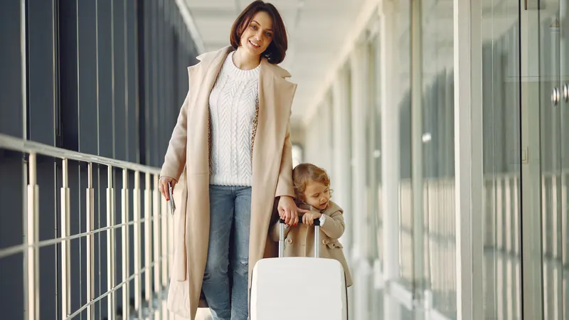 Mother and young daughter with a white suitcase walking along a glass-walled airport corridor