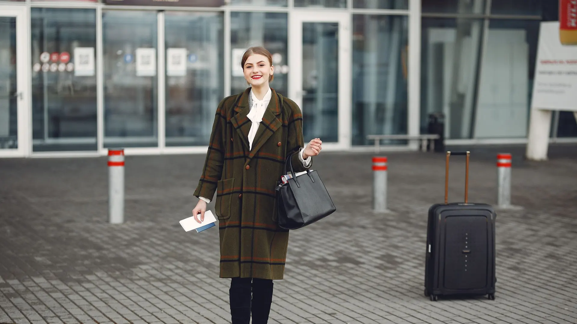 Woman in a long coat holding a boarding pass and a handbag beside a black suitcase outside an airport terminal