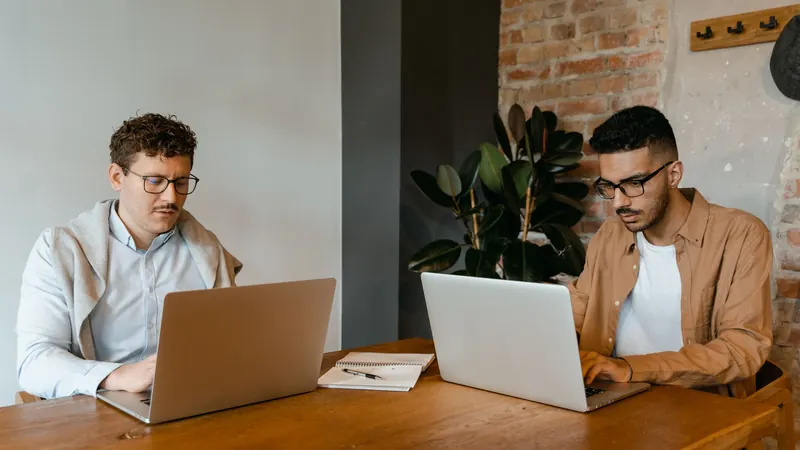 Two men working on laptops at a wooden table in a bright office with brick walls
