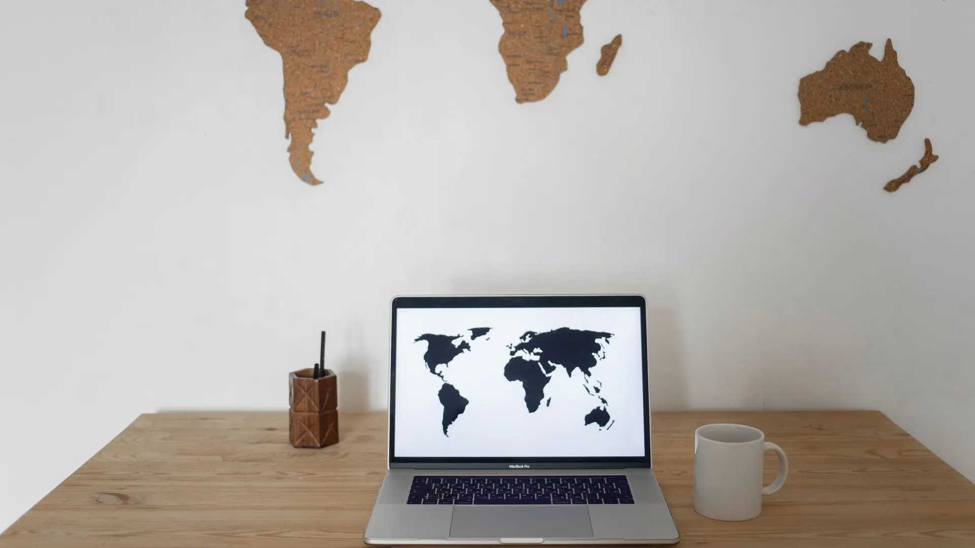 Laptop showing a world map on a wooden desk with a coffee cup, in front of a wall map